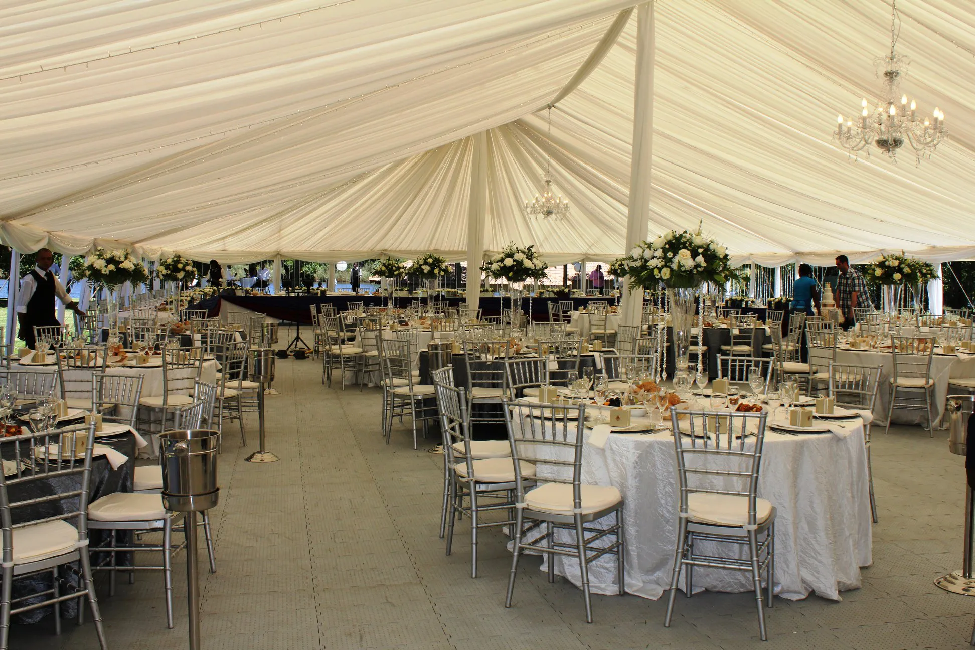 A draped marquee set with round tables, silver chairs and chandeliers